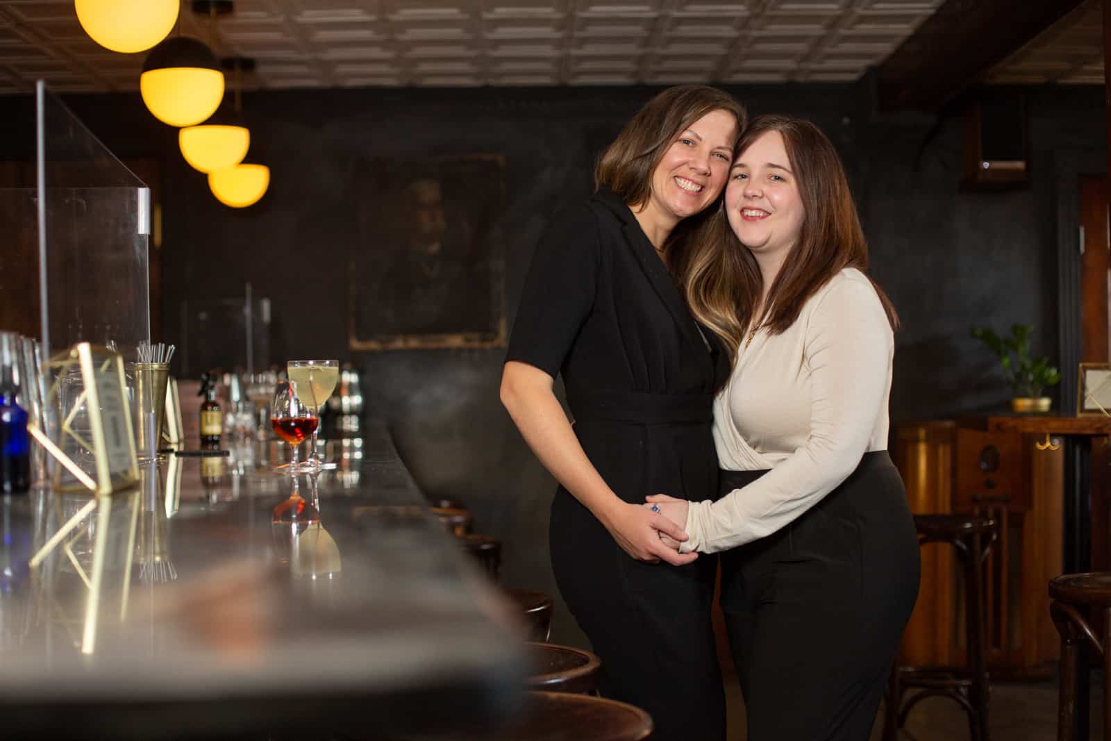 Two women hug standing next to a dimly lit cocktail bar